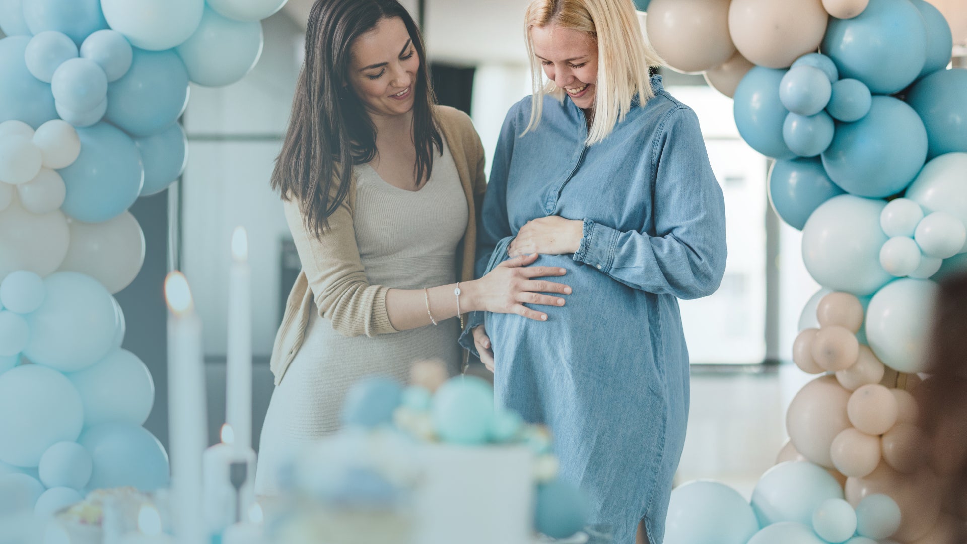 Two women standing in a room decorated with blue and white balloons, one of whom is pregnant.